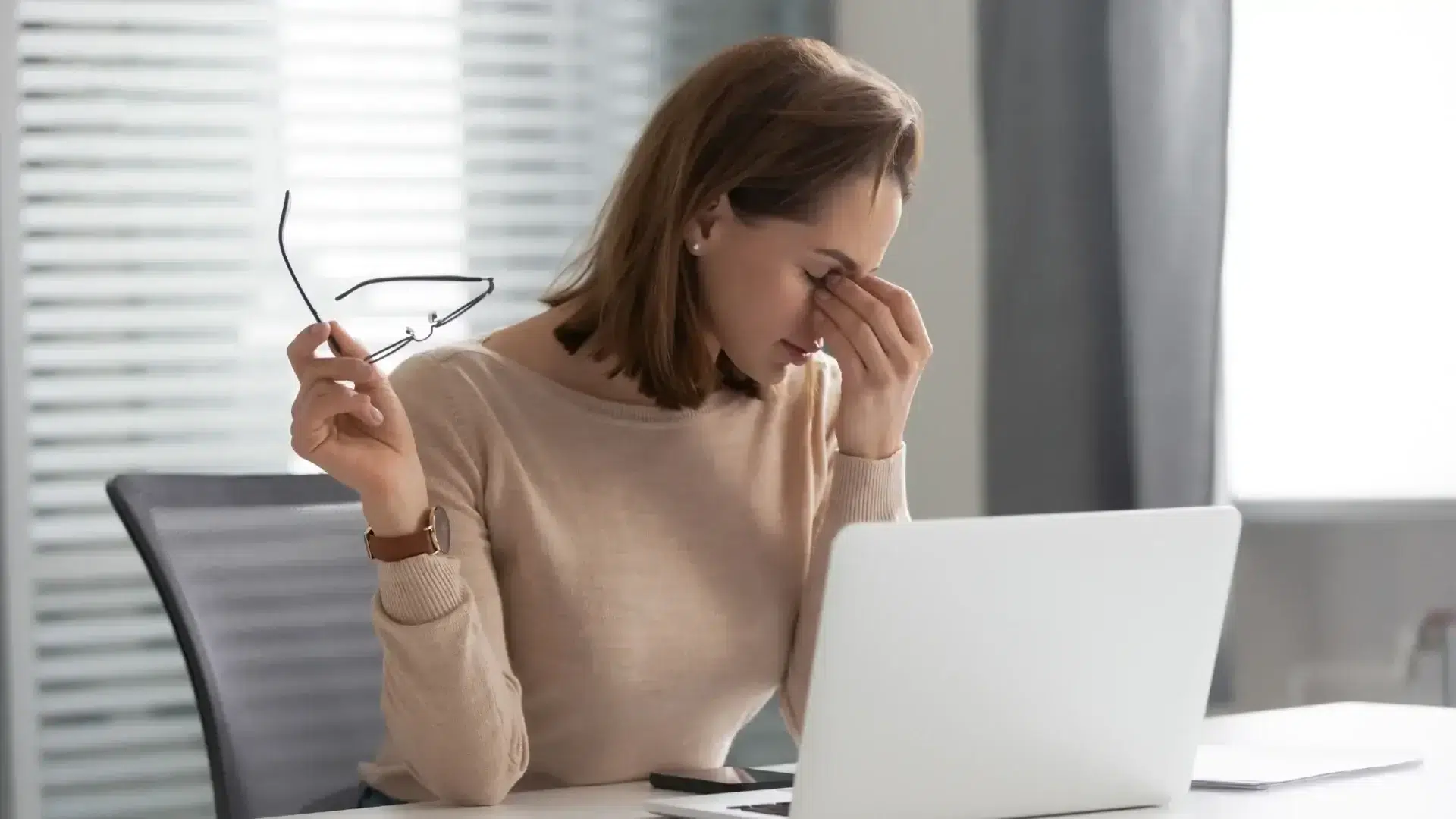 Exhausted woman holding glasses and pinching nose bridge, depicting common brain fog symptoms.